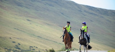 Two horses and riders on narrow road with upland hillside behind in gentle sunshine