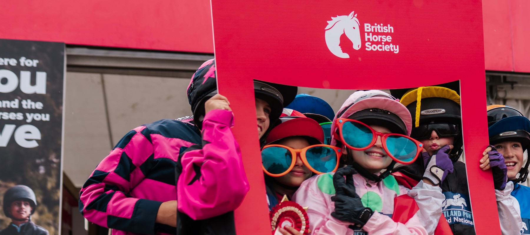 Children in jockey silks with large comedy coloured sunglasses, smiling and posing with a BHS card photo frame
