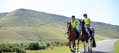 Two horses and riders on narrow road with upland hillside behind in gentle sunshine