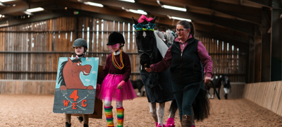Two children in fancy dress walking alongside a horse being led by an adult in an inside arena
